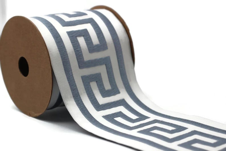 May include: A spool of white ribbon featuring a repeating gray Greek key pattern. The ribbon is wound around a circular brown cardboard core, with the patterned fabric extending outward to display the geometric design clearly.