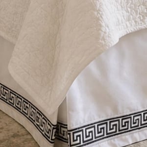 May include: White bed skirt with a black Greek key border. The bed skirt is paired with a white quilted bedspread. The bedding is on a neutral-toned rug.