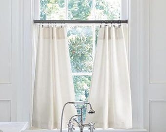 Puede incluir: Cortinas de ventana cortas de color crema cuelgan frente a una ventana con vista a árboles verdes. Las cortinas están colgadas de una barra de metal oscuro. Un grifo y un cabezal de ducha cromados están frente a la ventana.