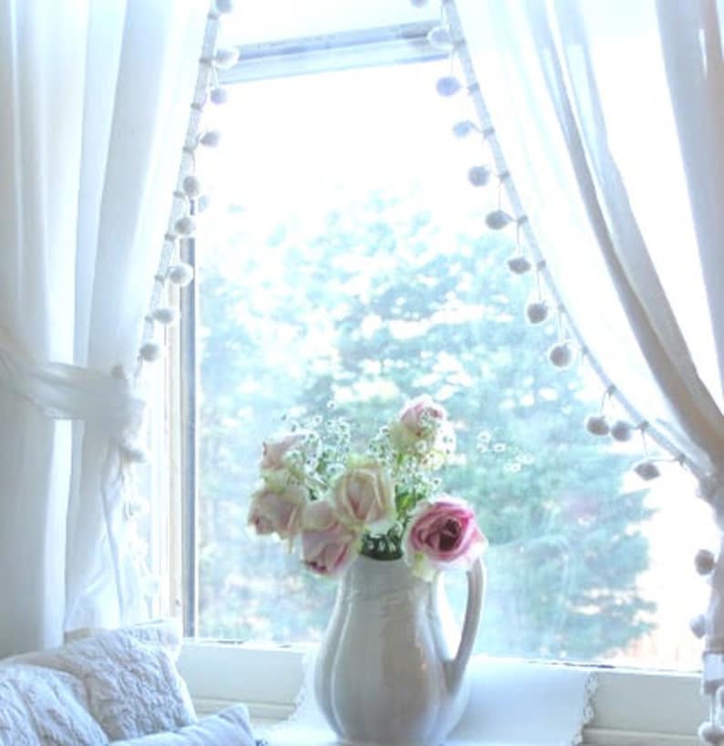 May include: White curtains with pom-pom trim frame a window with a view of trees. A white pitcher filled with pink and white roses sits on a windowsill.