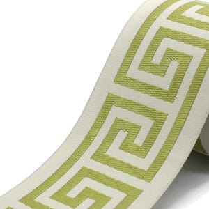 May include: A roll of woven ribbon featuring a repeating lime green Greek key pattern on a cream-colored background. The ribbon has a textured surface and is wound around a cardboard spool, displaying a continuous geometric design.