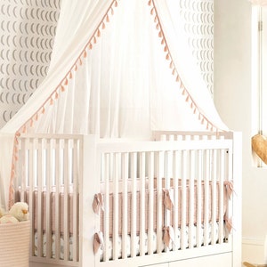 May include: A white wooden crib with a slatted design and a storage drawer at the base. The crib features a soft pink quilted bumper pad, a patterned mattress sheet, and a sheer white canopy overhead with delicate pink tassel trim accents.