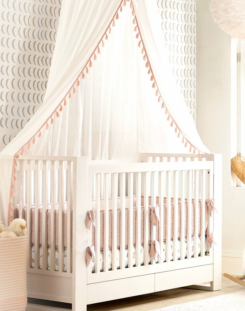 May include: A white wooden crib with a slatted design and a storage drawer at the base. The crib features a soft pink quilted bumper pad, a patterned mattress sheet, and a sheer white canopy overhead with delicate pink tassel trim accents.
