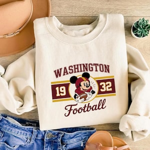 Puede incluir: Sudadera color crema con un dibujo animado de Mickey Mouse con casco de fútbol y balón. La sudadera tiene el texto "WASHINGTON" encima del dibujo, con "19" y "32" a cada lado y "Football" debajo.