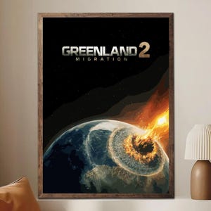 May include: A framed print featuring the text "GREENLAND 2 MIGRATION" above an illustration of Earth being struck by a fiery object. The artwork has a dark background with stars and a brown wooden frame.