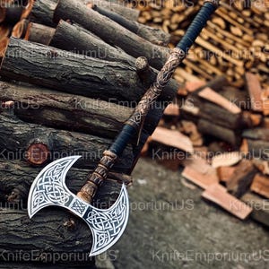 May include: A double-bladed axe with a silver blade and intricate Celtic knotwork design. The handle features a black grip and ornate bronze-colored detailing. The axe is displayed against a backdrop of stacked firewood, showcasing its craftsmanship.