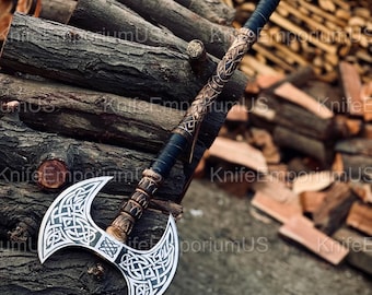 May include: A double-bladed axe with a silver blade and intricate Celtic knotwork design. The handle features a black grip and ornate bronze-colored detailing. The axe is displayed against a backdrop of stacked firewood, showcasing its craftsmanship.