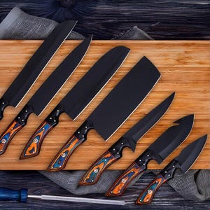 May include: A collection of seven black kitchen knives with patterned wooden handles in shades of orange, blue, and brown. The knives are displayed on a wooden board with a gray cloth and a sharpening steel with a blue handle.
