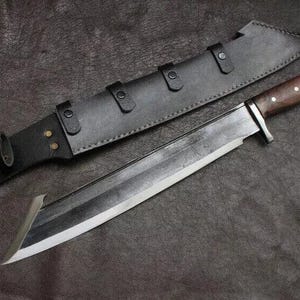 May include: A large, silver-bladed knife with a dark wooden handle and a black leather sheath. The knife has a wide blade and a curved tip. The sheath has three straps with buckles.