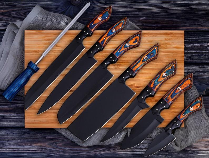May include: A collection of six black kitchen knives with decorative wooden handles in shades of blue, orange, and white. The knives are displayed on a wooden board with a sharpening steel. The blades are black.