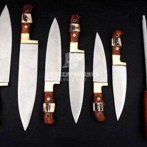 Può includere: Un set di sei coltelli da cucina e un'acciarino su sfondo nero. I coltelli hanno lame in acciaio lucido, manici in legno e dettagli decorativi. L'acciarino ha un manico in legno.