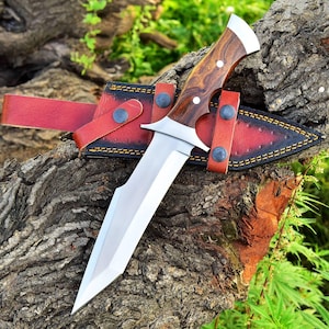 May include: A large hunting knife with a polished steel blade and a wooden handle. The knife is in a red leather sheath with black stitching and is secured with a strap. The knife is resting on a tree trunk.
