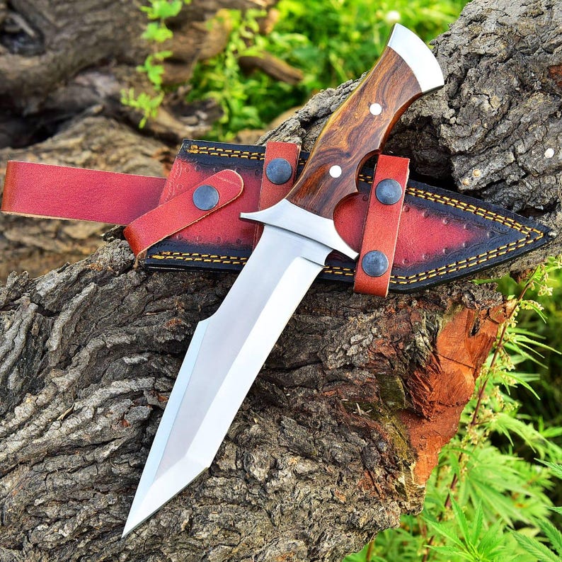 May include: A large hunting knife with a polished steel blade and a wooden handle. The knife is in a red leather sheath with black stitching and is secured with a strap. The knife is resting on a tree trunk.
