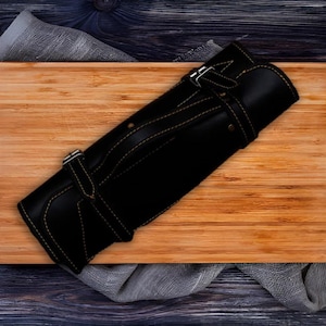 May include: Black leather knife roll with tan stitching and buckle fastenings. The roll is closed and rolled up, ready for storage or transport. The knife roll is on a wooden chopping board.