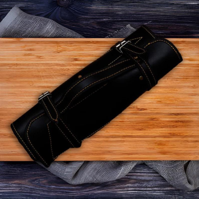 May include: Black leather knife roll with tan stitching and buckle fastenings. The roll is closed and rolled up, ready for storage or transport. The knife roll is on a wooden chopping board.