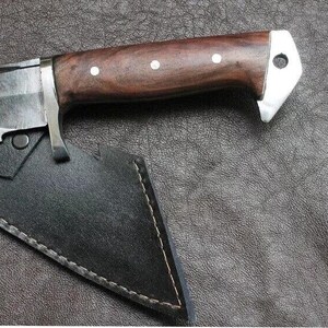 May include: Close-up of a hunting knife with a dark wooden handle and a silver bolster. The blade is partially visible, and the knife rests in a black leather sheath with white stitching. The handle has three white dot inlays.