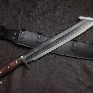 May include: A large, silver-bladed knife with a dark wooden handle and a black leather sheath. The knife has a unique, angled blade design and is resting on a textured, dark surface. The sheath has a belt loop.