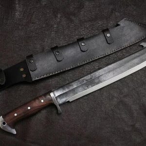 May include: A large, silver-bladed knife with a dark wooden handle and a black leather sheath. The knife has a wide blade and a sturdy design, suitable for outdoor or survival use. The sheath features multiple straps for secure storage.