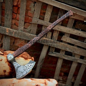 May include: A handcrafted Viking axe with a wooden handle featuring intricate carvings. The axe head is silver with a detailed Celtic knot design and a circular emblem. The axe is displayed against a rustic wooden backdrop.