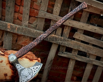 May include: A handcrafted Viking axe with a wooden handle featuring intricate carvings. The axe head is silver with a detailed Celtic knot design and a circular emblem. The axe is displayed against a rustic wooden backdrop.