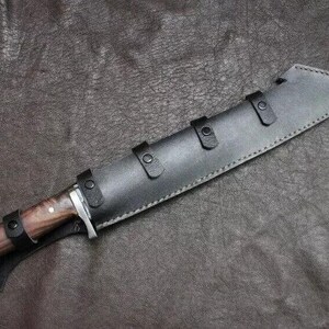 May include: A large hunting knife with a wooden handle and a black leather sheath. The knife blade is partially visible, and the sheath has three straps with buckles. The handle has a silver bolster.