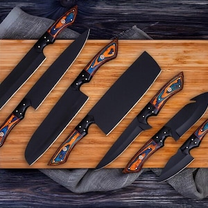 May include: A set of black kitchen knives with decorative wooden handles in orange, blue, and white. The knives are displayed on a wooden chopping board, with a grey cloth and a blue and silver tool in the background.