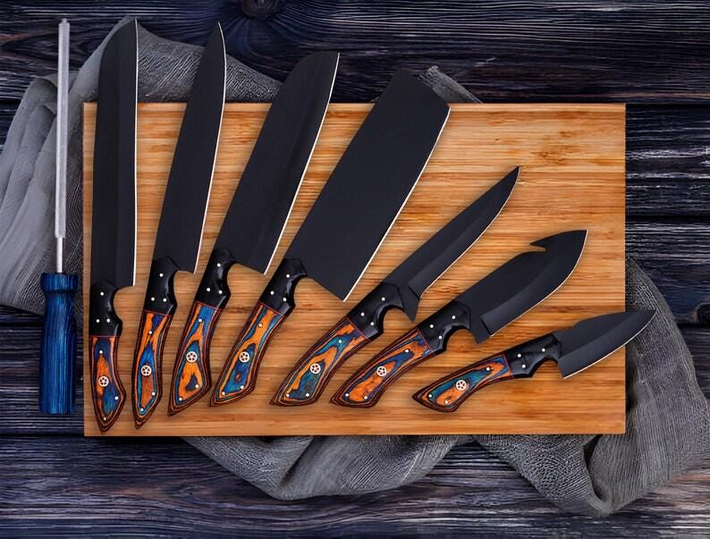 May include: A collection of seven black kitchen knives with patterned handles in blue, orange, and brown, displayed on a wooden chopping board. Includes a sharpening steel with a blue handle. Knives vary in size and shape.