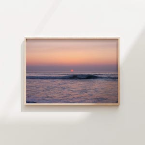 Puede incluir: Fotografía enmarcada de una puesta de sol sobre el océano. El cielo transita del rosa al naranja, con un sol rojo que se pone sobre el agua. Se ven olas en primer plano y el marco es de color madera clara.