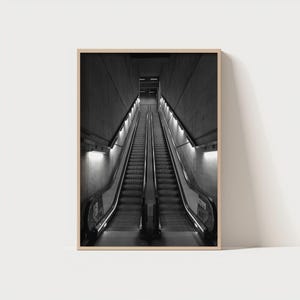 Può includere: Fotografia in bianco e nero di due scale mobili che salgono in un tunnel di cemento scuro. Le scale mobili sono parallele, con corrimano e luci ai lati. L'immagine è incorniciata in una cornice di legno chiaro.
