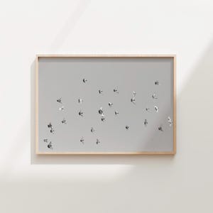Puede incluir: Una impresión enmarcada que presenta una bandada de pájaros en vuelo sobre un fondo gris apagado. Las aves se representan en tonos grises y blancos, creando una sensación de movimiento. El marco es de color madera clara.