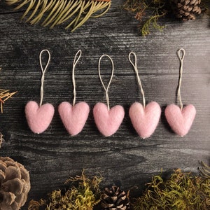 May include: Five pink felt heart ornaments with white string loops, arranged on a dark wood surface. The hearts are surrounded by pinecones, evergreen sprigs, and moss, creating a rustic, seasonal theme.