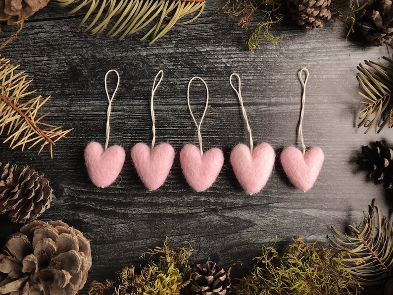 May include: Five pink felt heart ornaments with white string loops, arranged on a dark wood surface. The hearts are surrounded by pinecones, evergreen sprigs, and moss, creating a rustic, seasonal theme.