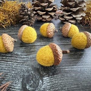May include: A collection of handmade felt acorns with brown caps and yellow bodies. The acorns are arranged on a dark wooden surface, with pine cones and dried foliage in the background. The acorns are a fall-themed decorative item.