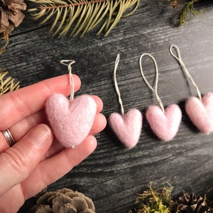 May include: A collection of small, light pink felt heart ornaments with silver string loops for hanging. The hearts are displayed against a dark wood background with natural elements like moss and pinecones, creating a rustic aesthetic.