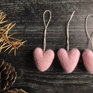 May include: Close-up of three pink felt heart ornaments with string loops, arranged on a dark, textured surface. A sprig of dried pine needles and a pine cone are in the background. The hearts are soft and fluffy.
