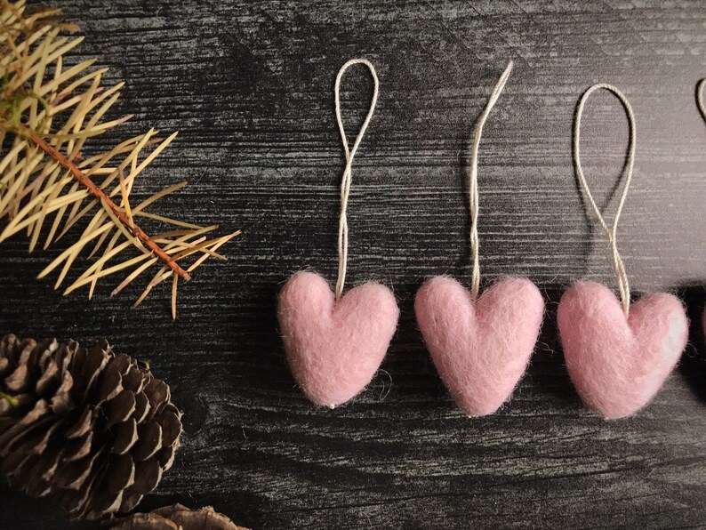 May include: Close-up of three pink felt heart ornaments with string loops, arranged on a dark, textured surface. A sprig of dried pine needles and a pine cone are in the background. The hearts are soft and fluffy.