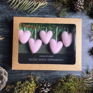 May include: A box of pink felted wool heart ornaments. The ornaments are displayed in a brown cardboard box with a clear window. The box is surrounded by pine needles, moss, and pine cones. The text "HOUSE OF MOSS" and "FELTED WOOL ORNAMENTS" is visible.