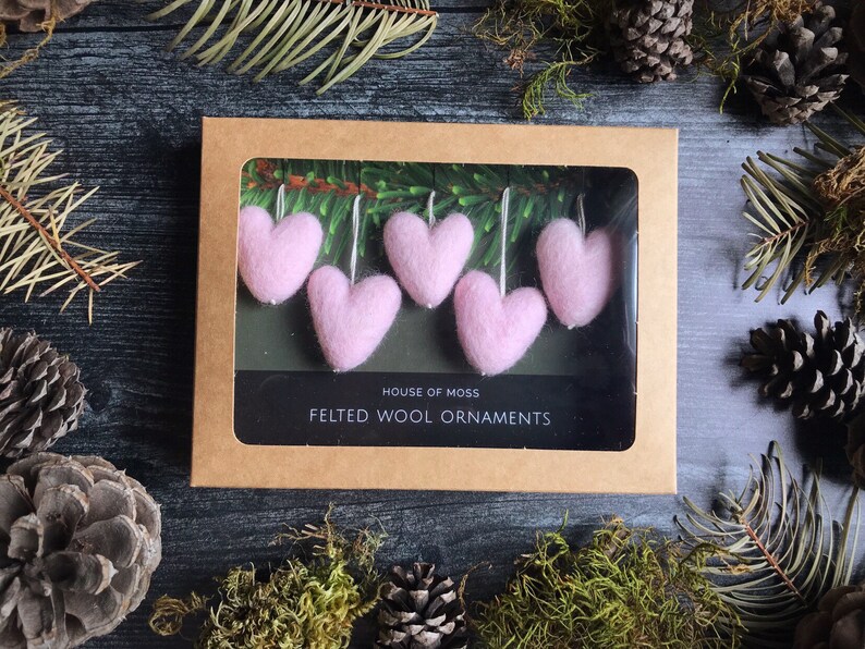 May include: A box of pink felted wool heart ornaments. The ornaments are displayed in a brown cardboard box with a clear window. The box is surrounded by pine needles, moss, and pine cones. The text "HOUSE OF MOSS" and "FELTED WOOL ORNAMENTS" is visible.