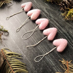 May include: Five light pink felt heart ornaments with string loops are arranged diagonally on a dark wood surface. The hearts are surrounded by pine cones, moss, and greenery, creating a rustic, natural aesthetic. These ornaments are ideal for Valentine's Day or other special occasions.