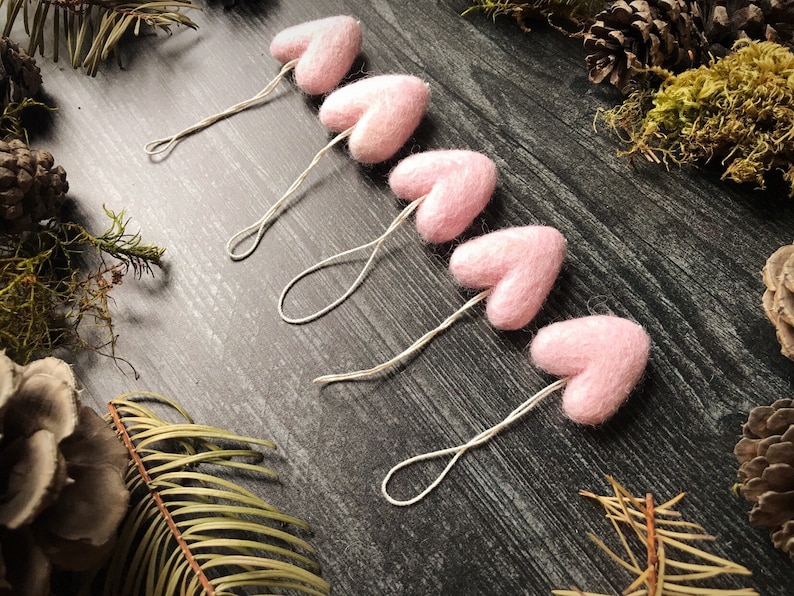 May include: Five light pink felt heart ornaments with string loops are arranged diagonally on a dark wood surface. The hearts are surrounded by pine cones, moss, and greenery, creating a rustic, natural aesthetic. These ornaments are ideal for Valentine's Day or other special occasions.