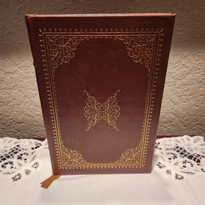 May include: A brown leather-bound book with gold filigree detailing on the cover and spine. The cover features a gold butterfly design in the center. A brown ribbon bookmark is visible.