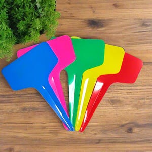 May include: A set of five T-shaped plant markers in vibrant colors including blue, pink, green, yellow, and red. Each marker features a flat rectangular top section for labeling and a pointed stake base designed for insertion into soil or potting mix.