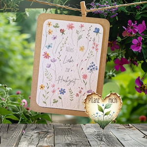 Könnte beinhalten: Eine florale Hochzeitskarte mit dem Text "Alles Liebe zur Hochzeit" und einem herzförmigen Anhänger mit der Aufschrift "Pflanz mich!". Die Karte ist mit Aquarellblumen in verschiedenen Farben verziert und hängt an einer Wäscheleine.