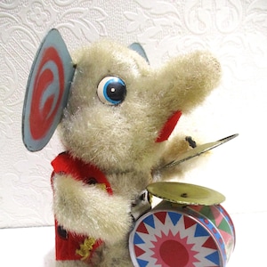 May include: Vintage wind-up toy elephant with a red vest, blue and red ears, and a drum. The elephant is playing cymbals and has a drum with a red and white starburst design. The toy is made of plush material.