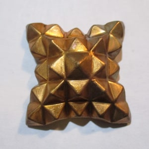 May include: A gold-colored, geometric decorative object with a square shape and a repeating pyramid pattern. The object has a textured surface and a metallic sheen, suggesting it is made of metal or a similar material. The design is symmetrical and detailed.
