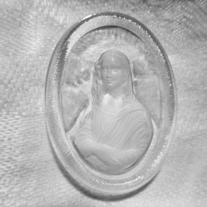 May include: A clear, oval-shaped glass cameo featuring a bas-relief portrait of a woman with her arms crossed. The portrait is framed by a textured border. The cameo is set against a soft, textured background.