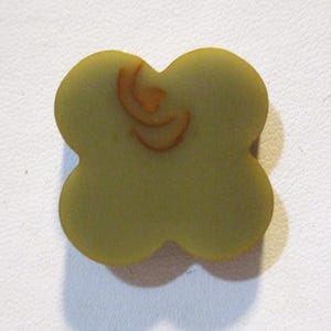 May include: A four-leaf clover-shaped object in a muted olive green color. The clover has a slightly textured surface and a small, abstract, orange-brown design on one of the leaves. The background is a plain white surface.