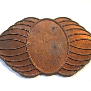 Peut inclure: Un emblème en métal cuivré vintage avec un centre ovale et des formes en forme d'ailes stylisées. La surface est texturée et présente une apparence légèrement vieillie, suggérant qu'il s'agit d'une pièce ancienne ou historique.