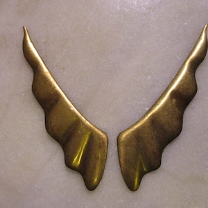 May include: A pair of gold-colored metal wings with a textured, wavy design. The wings are symmetrical and have a pointed tip.