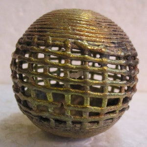 May include: A spherical, decorative object with a cage-like design. The exterior is a metallic gold color with a textured, woven appearance. The sphere is approximately 3 inches in diameter, with a dark, aged patina.